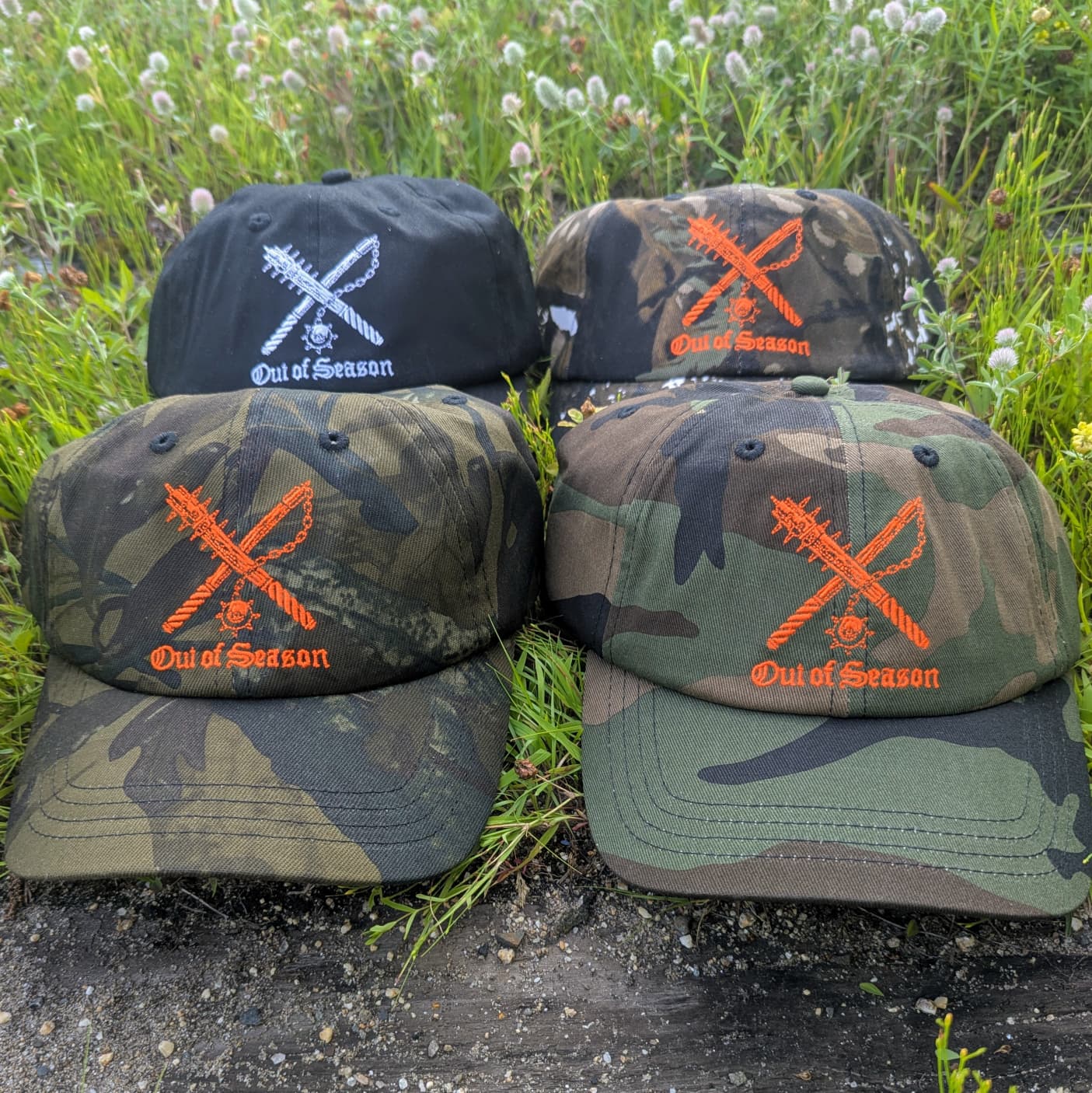 Four baseball caps arranged outdoors on grass and dirt, featuring camouflage and solid color designs with orange embroidered crossed tools and the text 'Out of Season'.