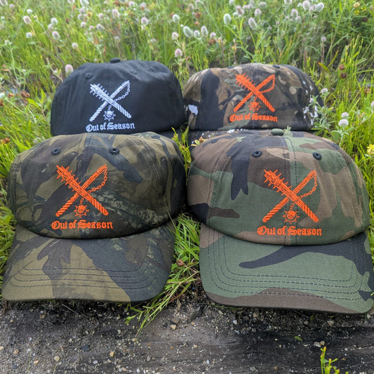 Four baseball caps arranged outdoors on grass and dirt, featuring camouflage and solid color designs with orange embroidered crossed tools and the text 'Out of Season'.