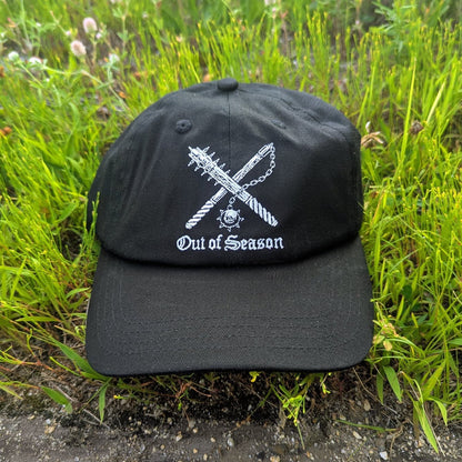 A black baseball cap with a white embroidered graphic of crossed hockey sticks and the text 'Out of Season' resting on green grass.