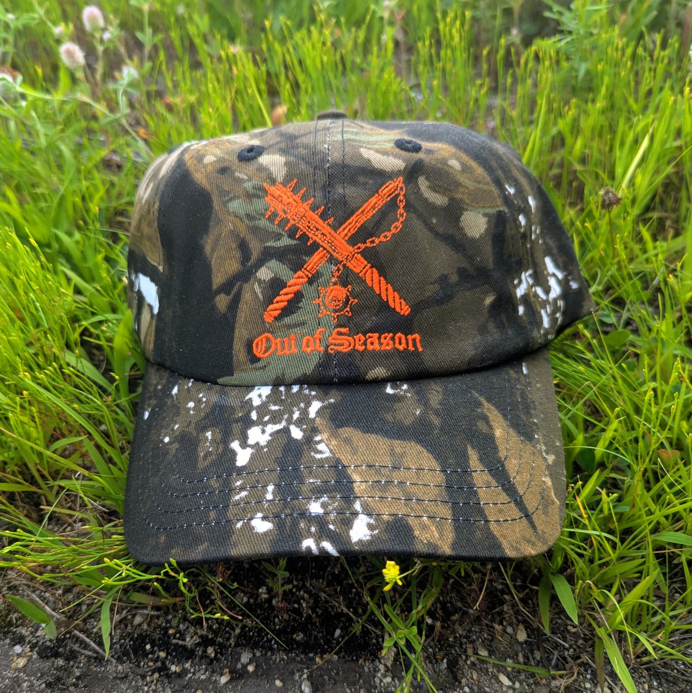 A camouflage baseball cap with an orange embroidered logo of crossed chainsaws and the text 'Out of Season' resting on green grass.