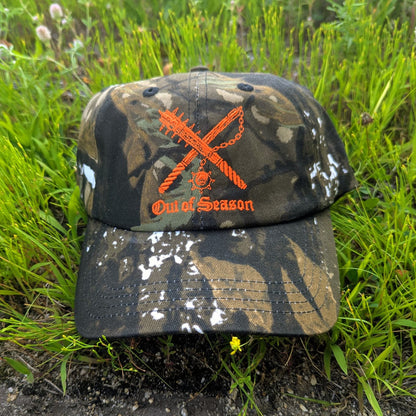 A camouflage baseball cap with an orange embroidered logo of crossed chainsaws and the text 'Out of Season' resting on green grass.