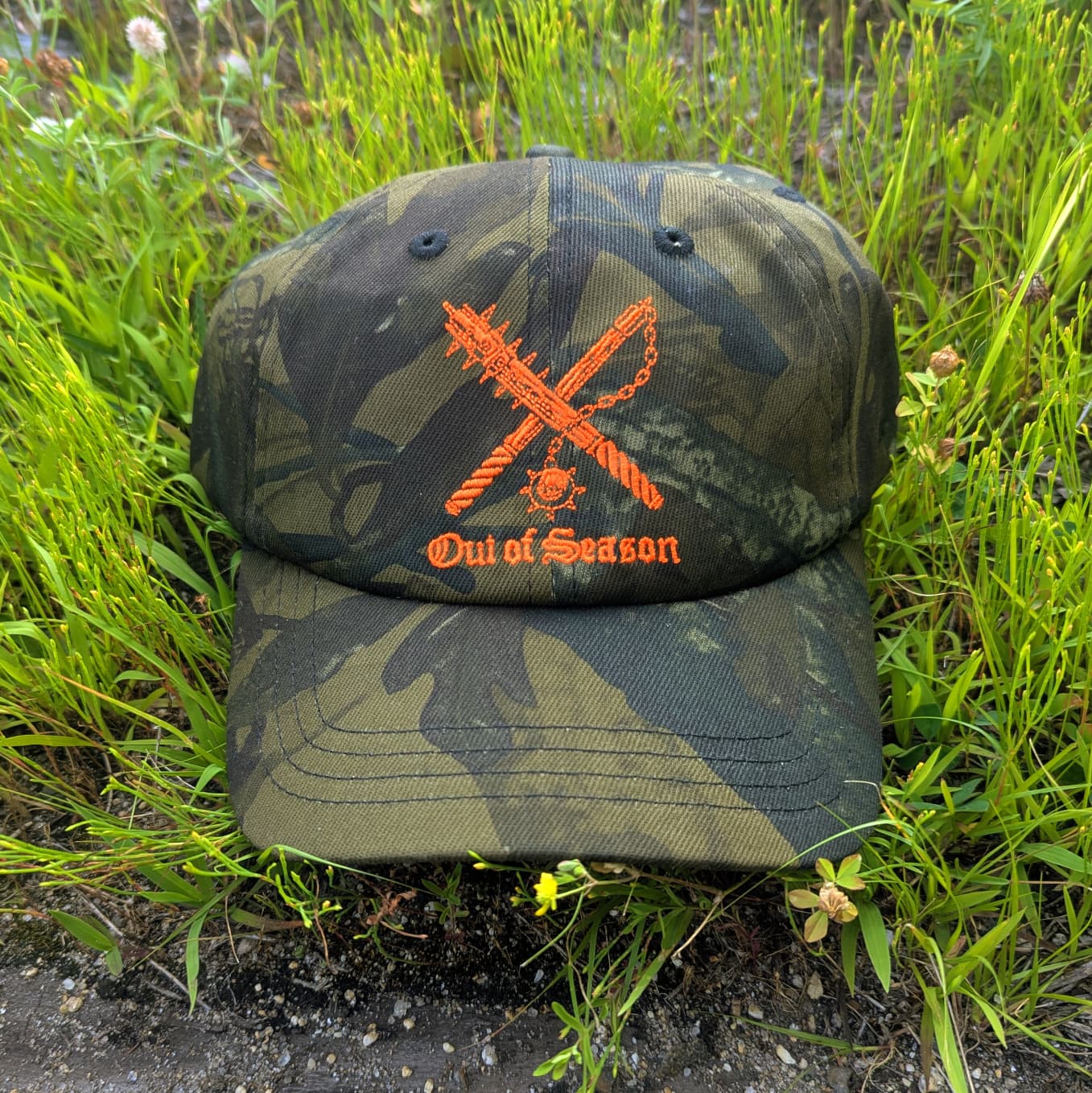 A camouflage baseball cap with an orange embroidered design featuring crossed chains and the text 'Out of Season,' placed on grass and dirt.