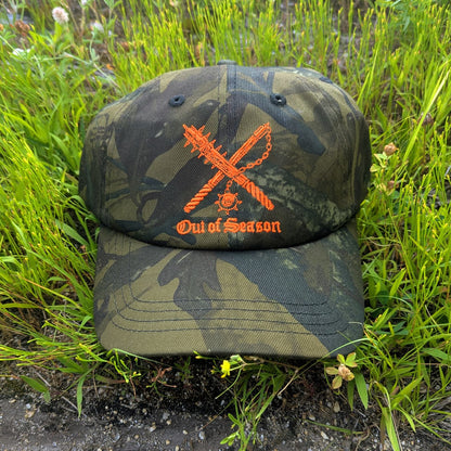 A camouflage baseball cap with an orange embroidered design featuring crossed chains and the text 'Out of Season,' placed on grass and dirt.