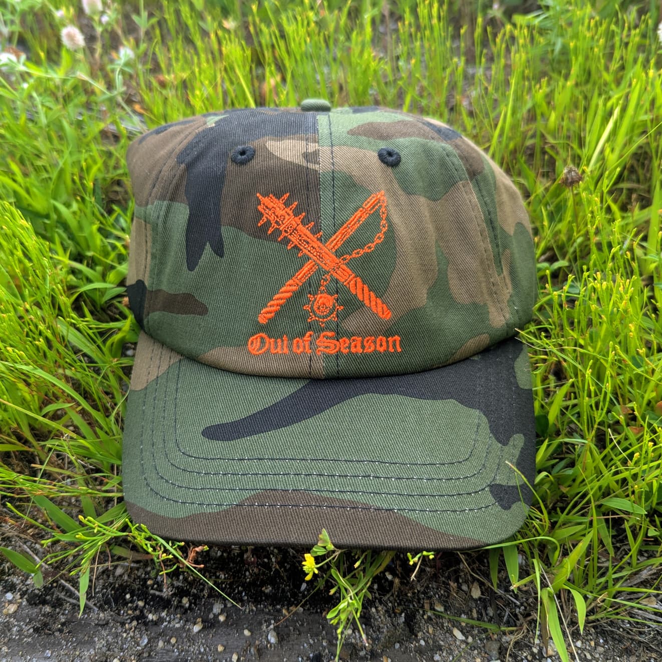 A camouflage baseball cap with an orange embroidered 'Out of Season' logo featuring crossed weapons, resting on green grass.