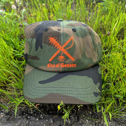 A camouflage baseball cap with an orange embroidered 'Out of Season' logo featuring crossed weapons, resting on green grass.