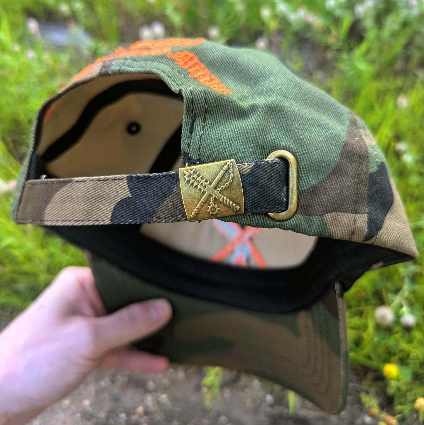 A camouflage baseball cap held by a hand outdoors, showing the back strap closure with a brass buckle and a gold-toned triangular emblem.