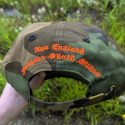 A camouflage baseball cap held by a hand outdoors, featuring orange embroidered text