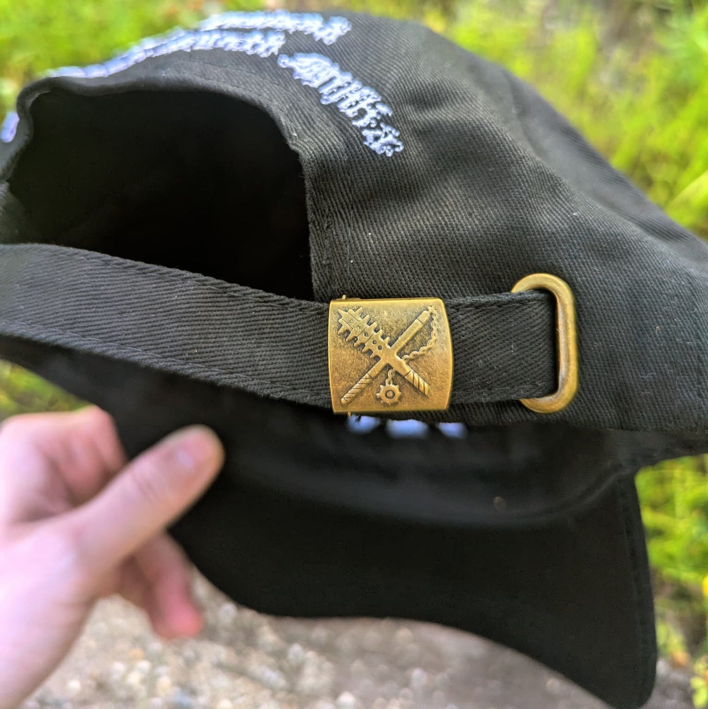A close-up of a black baseball cap being held by a hand, showing a black adjustable strap with a square gold-toned metal emblem and a gold-toned ring. The cap has white embroidered text on the side, and the background is a blurred outdoor setting with green grass and light-colored ground.