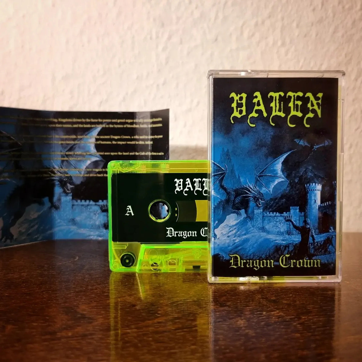 A cassette tape and its printed cover case displayed on a wooden surface against a textured wall. The cassette is translucent neon green with a black label, and the cover art shows a dark blue dragon over a castle with the text 'VALEN' and 'Dragon-Crown'.